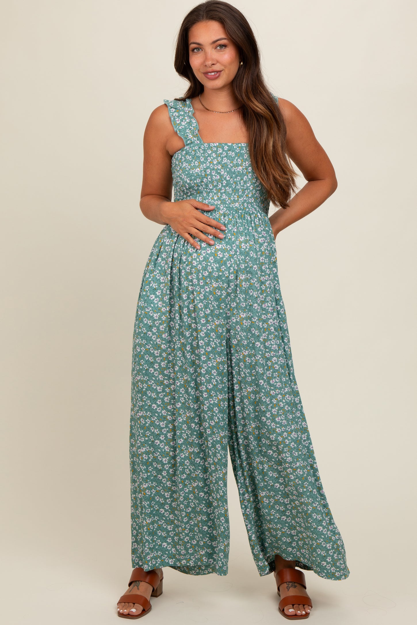 woman wearing a green floral maternity jumpsuit with a smocked bodice and one ruffled shoulder strap, showing her baby bump