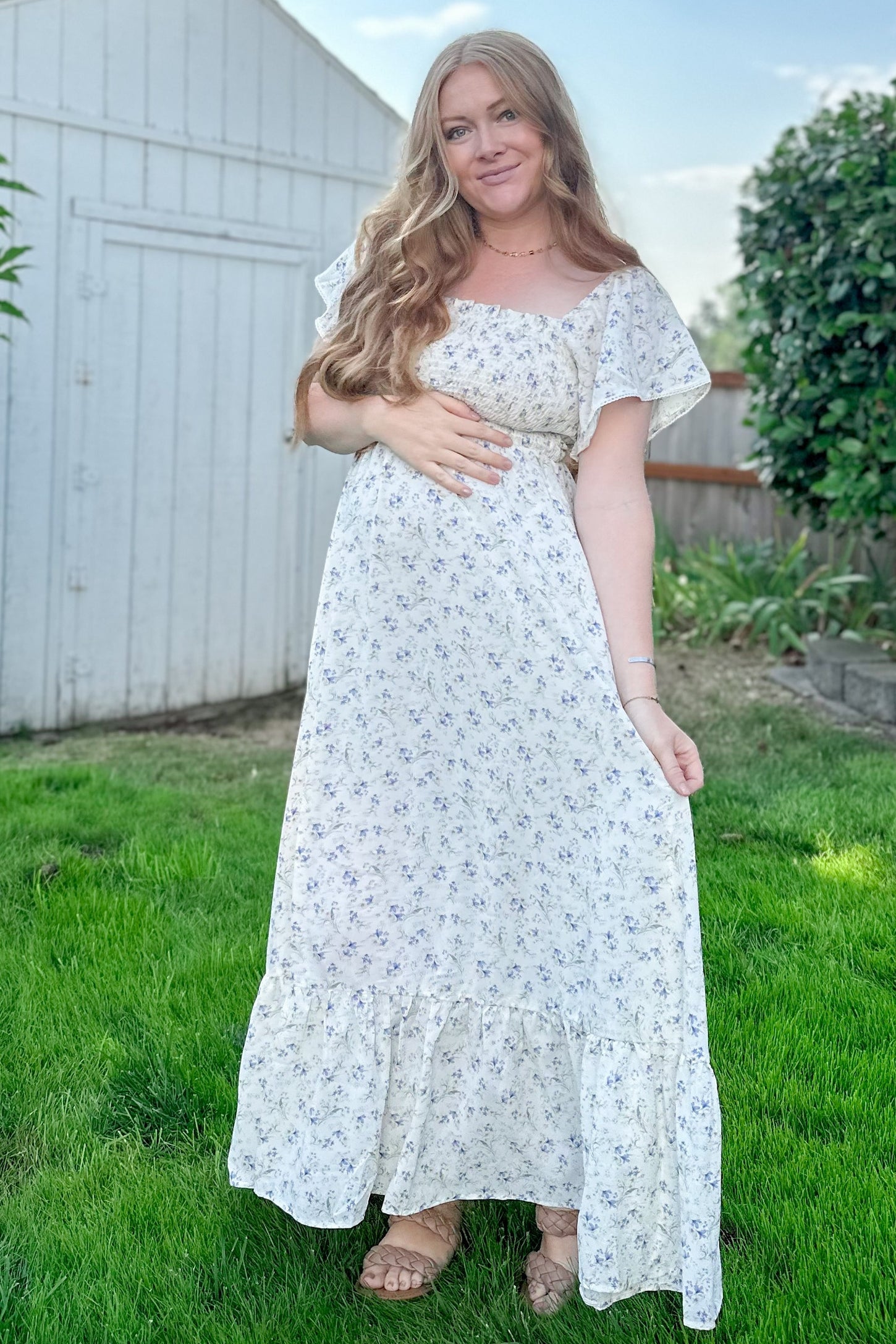 woman wearing a white floral maternity dress and holding her baby bump in a grassy outdoor setting