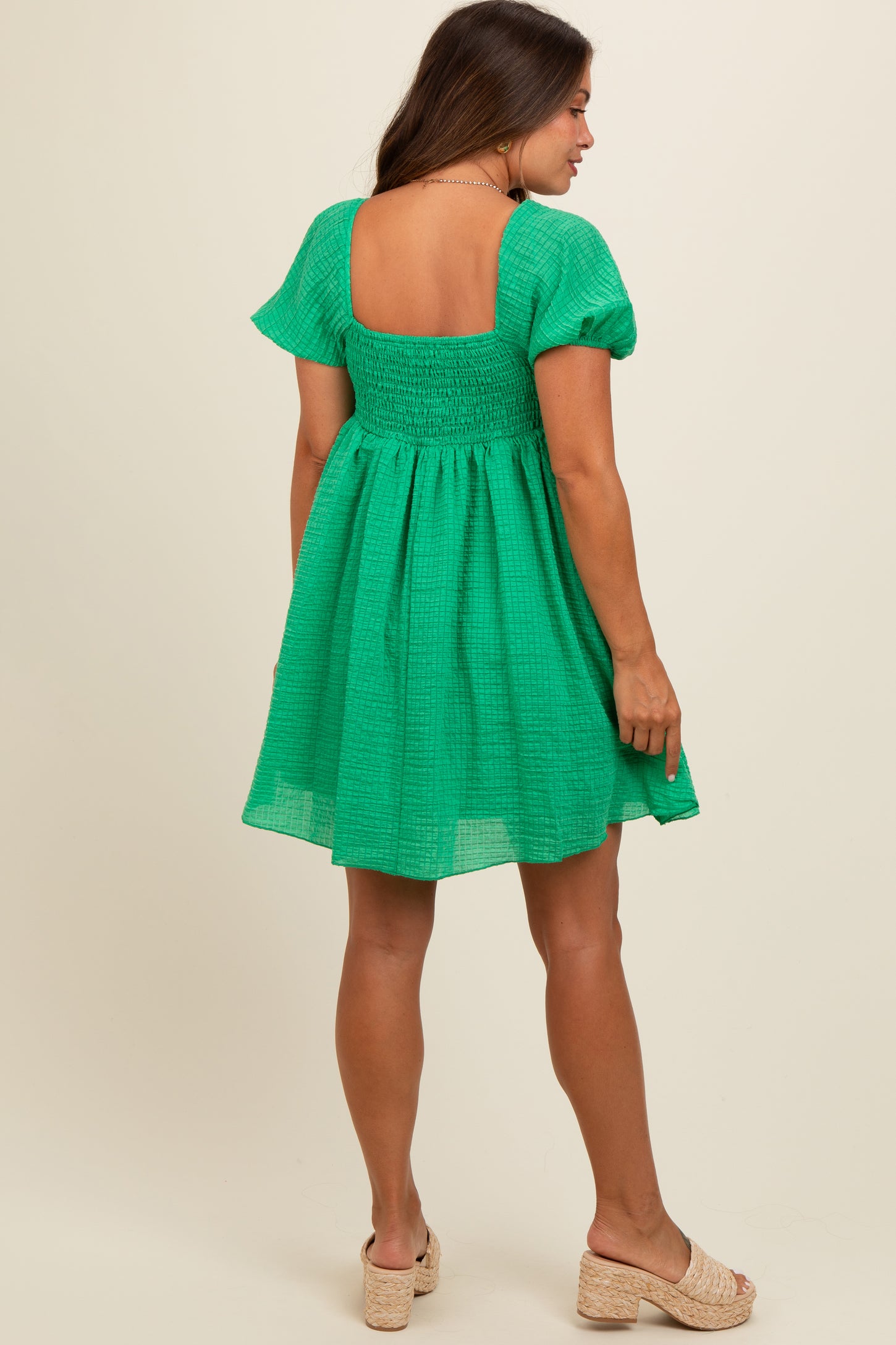 woman wearing a green smocked maternity dress with short sleeves and a square neckline, standing with her back to the camera