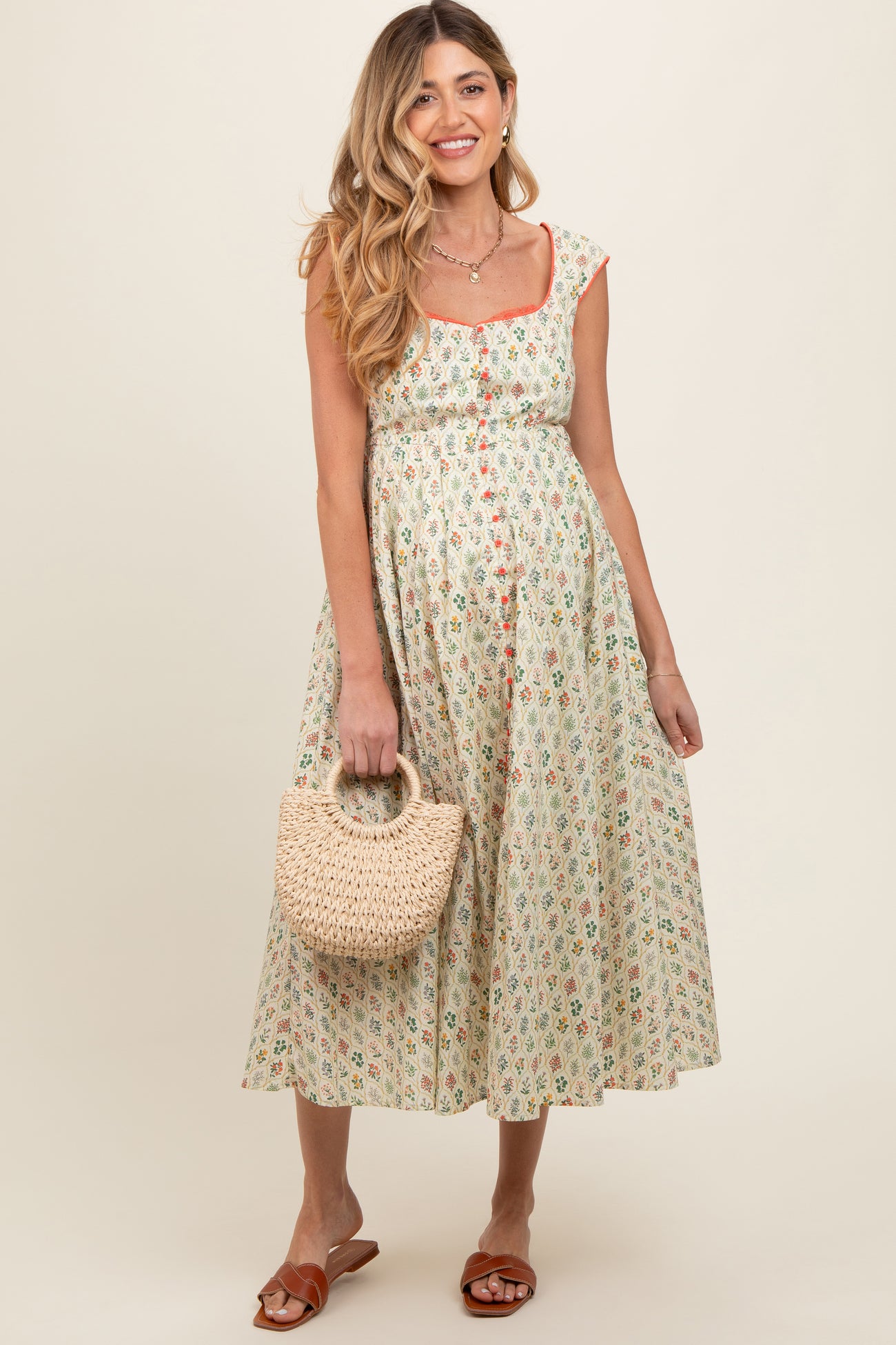 woman wearing a floral print maternity dress with a square neckline and carrying a woven handbag, showing her baby bump