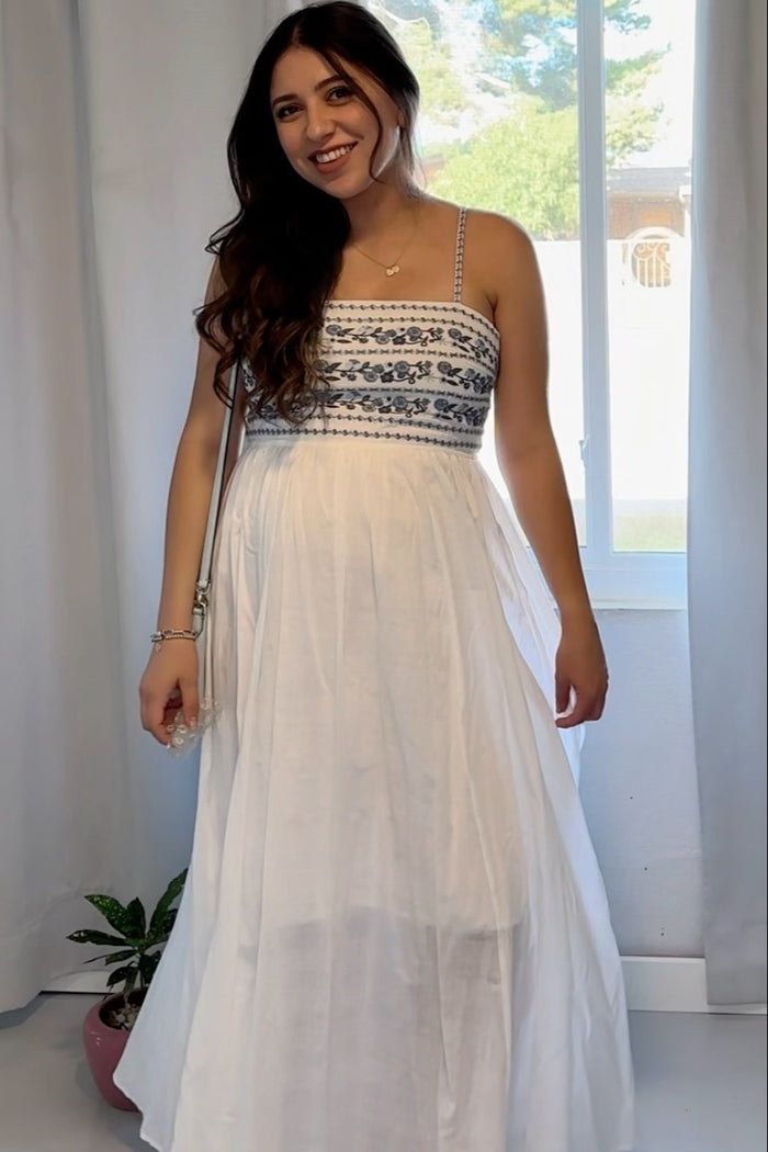 woman wearing a white maternity maxi dress with embroidery, showing a baby bump, standing by a window