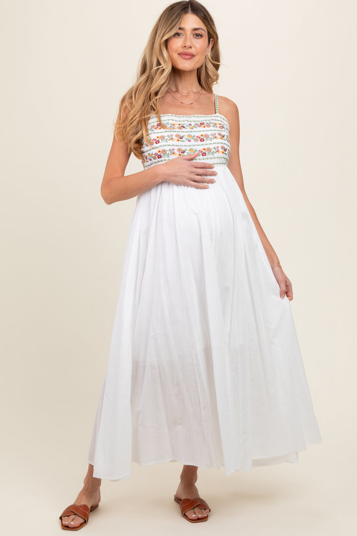 woman wearing a white maternity maxi dress with colorful embroidery and holding her baby bump