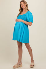 woman wearing a bright turquoise maternity dress with a textured bodice and puff sleeves, showcasing her baby bump