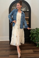woman wearing a white tiered maxi dress with floral details and a denim jacket, posed with one hand on hip