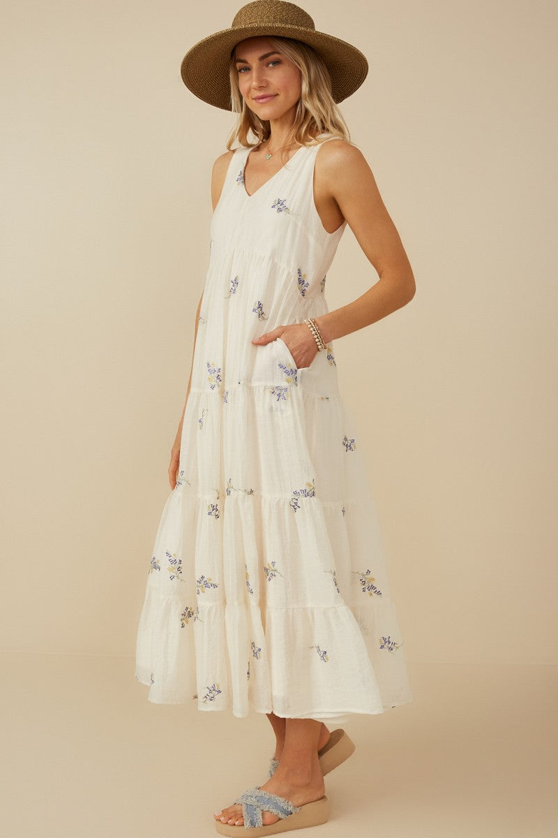woman wearing a white tiered maxi dress with floral embroidery and pockets, paired with a wide-brimmed hat and sandals