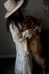 woman wearing a cream-colored cardigan and a white floral midi dress, holding a golden retriever in a cozy indoor setting