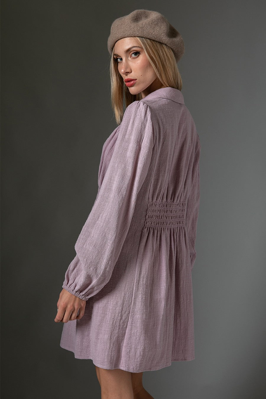 woman wearing a lilac long sleeve dress with a smocked waist and a beret, posing sideways against a gray background