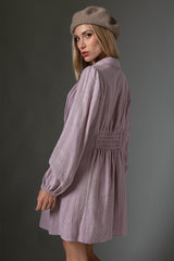 woman wearing a lilac long sleeve dress with a smocked waist and a beret, posing sideways against a gray background