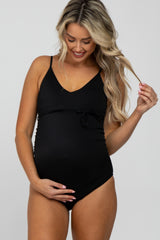 woman wearing a black ribbed maternity swimsuit with a tie detail, gently holding her baby bump