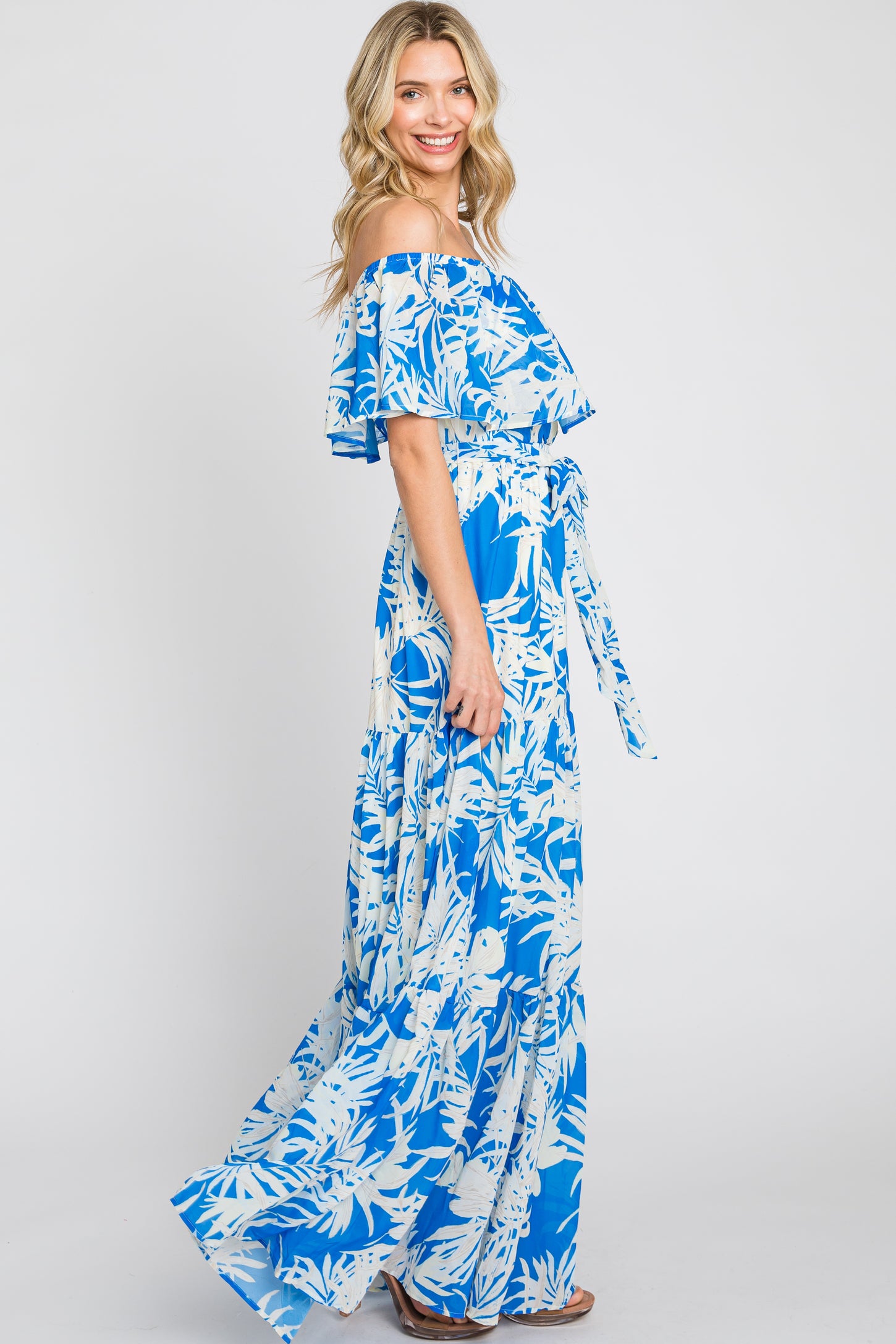 woman wearing a blue off-the-shoulder maxi dress with a white floral pattern and a tie waist detail