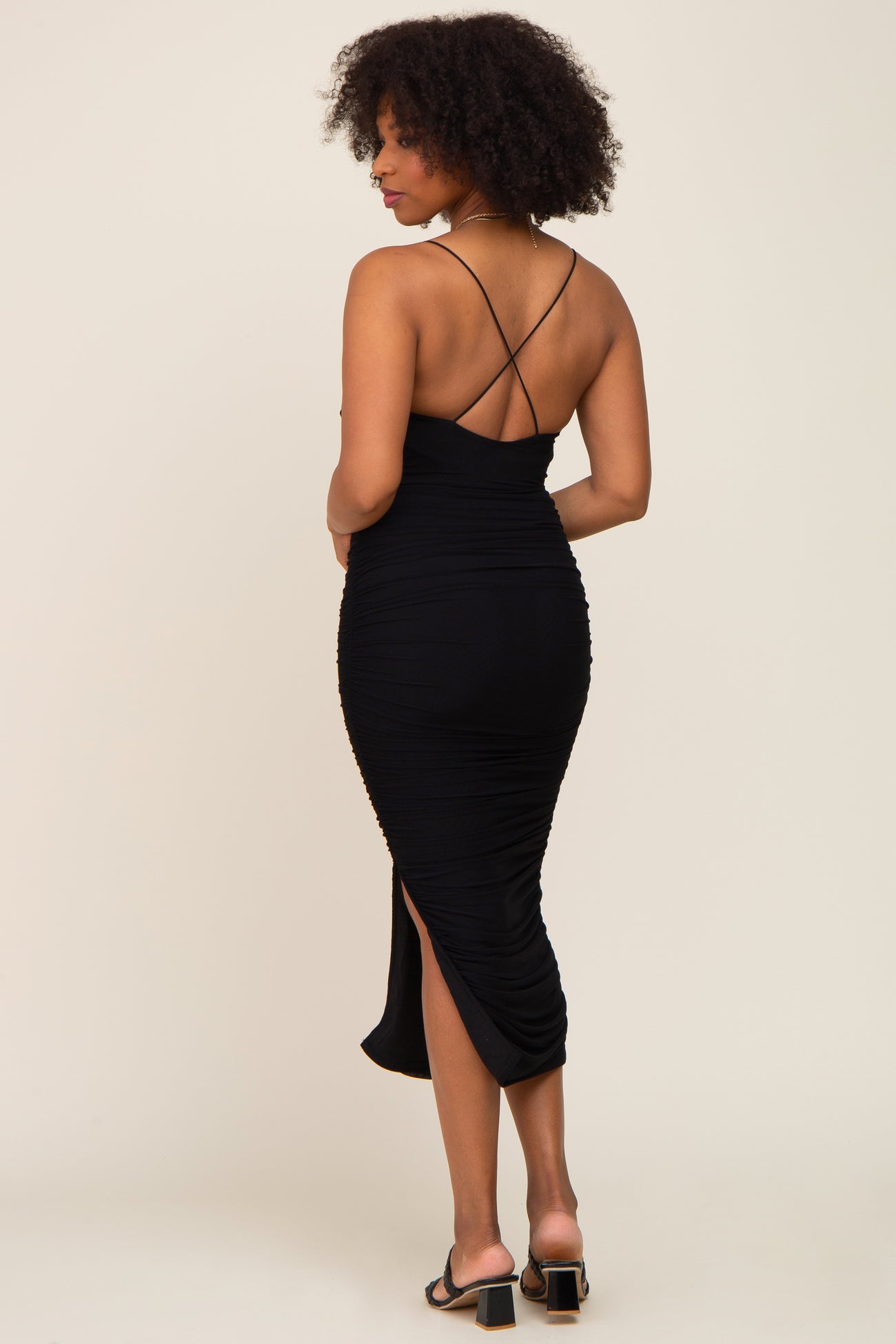 woman wearing a black ruched midi dress with a strappy back and side slit, standing sideways