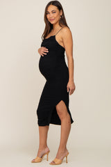 woman wearing a black ruched maternity dress with side slit, holding her baby bump and standing in heels