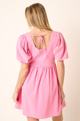 Back view of a woman wearing a pink puff sleeve mini dress with a square neckline and tie detail