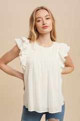 Woman wearing a white sleeveless blouse with eyelet ruffle shoulders and pleated bodice against a beige background