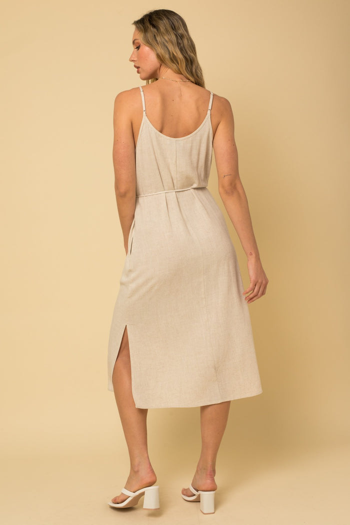 Back view of a woman wearing a beige linen midi dress with spaghetti straps and a side slit