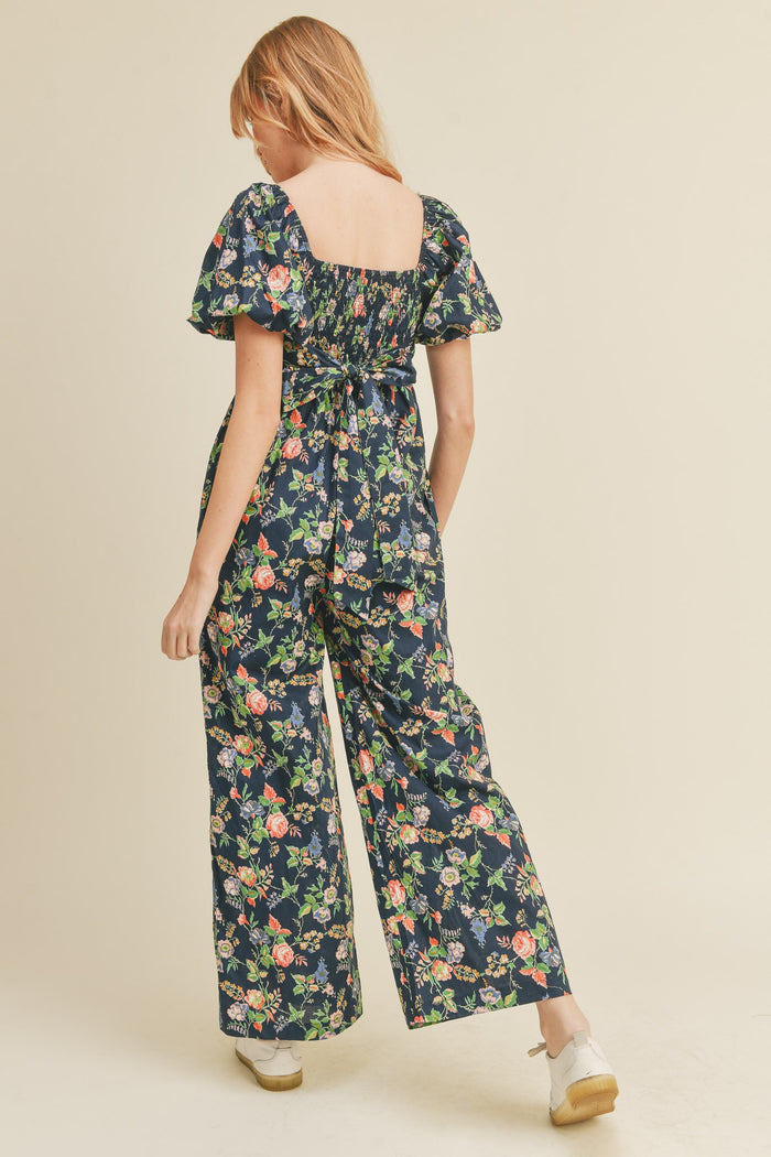 Back view of woman wearing navy floral wide leg jumpsuit with puff sleeves and smocked back detail