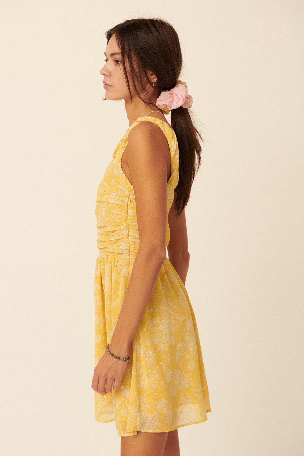 woman wearing a yellow sleeveless dress with a floral pattern and gathered details, standing sideways with hair in a ponytail