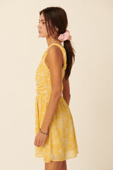 woman wearing a yellow sleeveless dress with a floral pattern and gathered details, standing sideways with hair in a ponytail