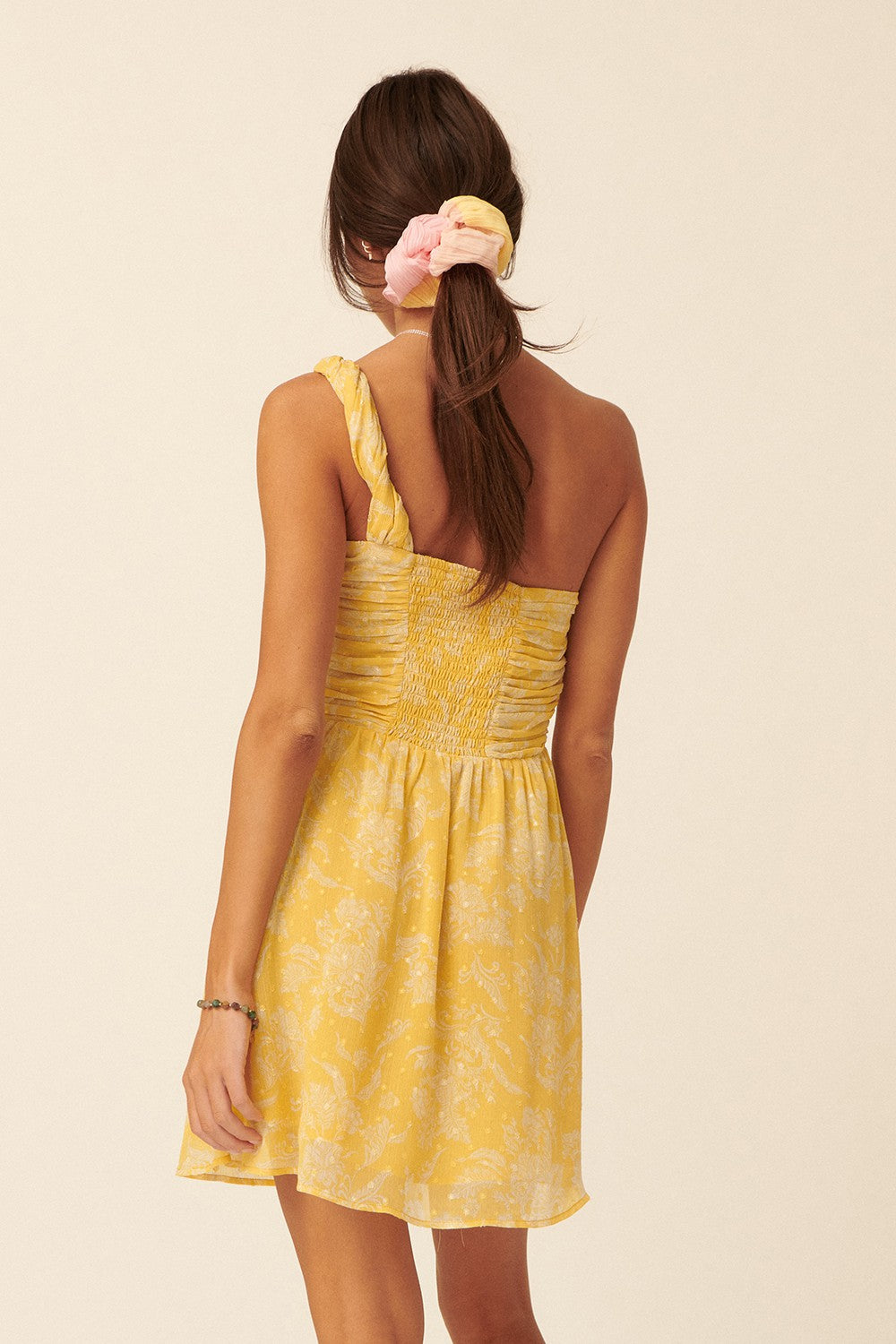 woman wearing a yellow floral print dress with a smocked bodice and one shoulder strap, viewed from the back