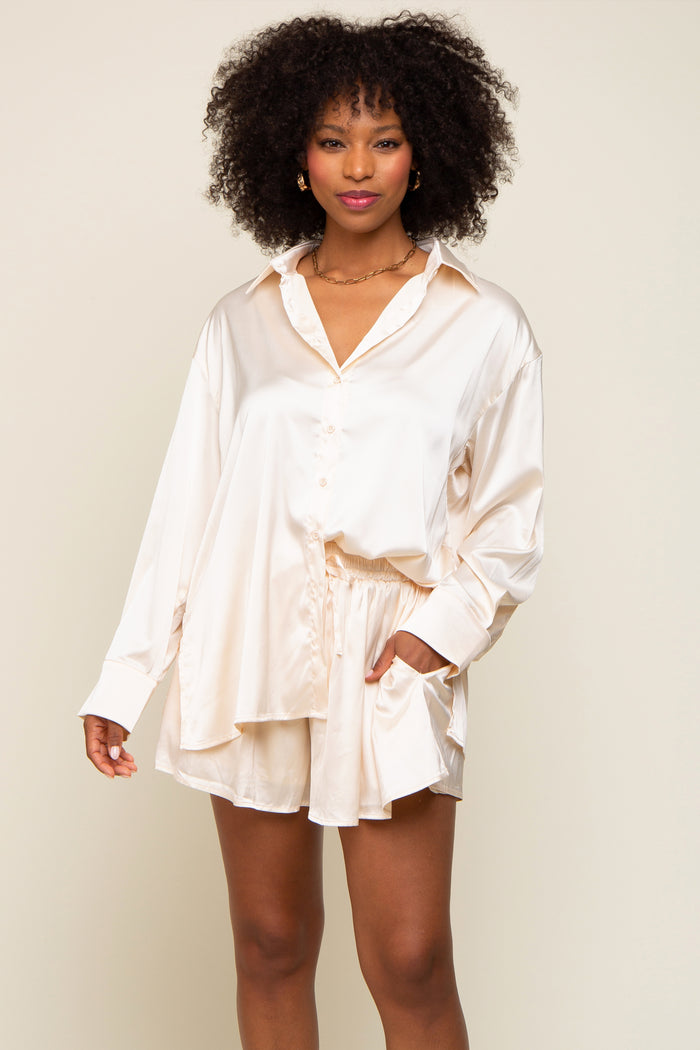 Woman wearing cream satin button-down shirt and matching shorts lounge set with gold chain necklace