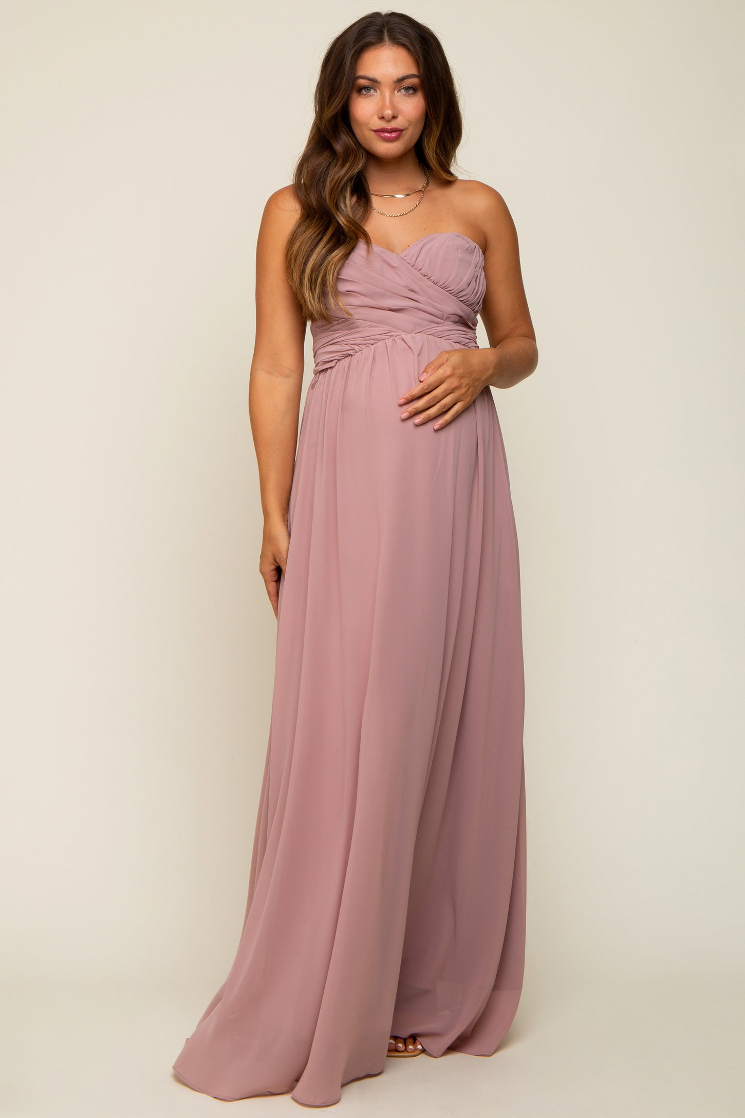 woman wearing a dusty rose strapless maternity gown with ruched details and holding her baby bump