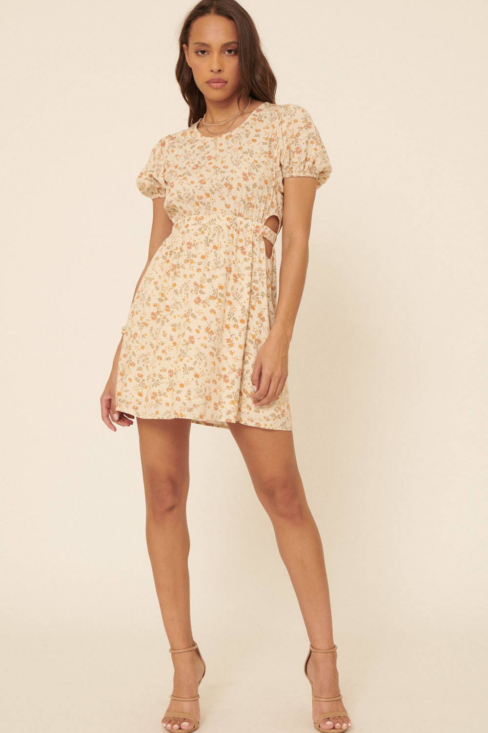 woman wearing a beige floral short dress with puff sleeves and cut-out sides, standing in a relaxed pose