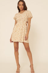 woman wearing a beige floral short dress with puff sleeves and cut-out sides, standing in a relaxed pose