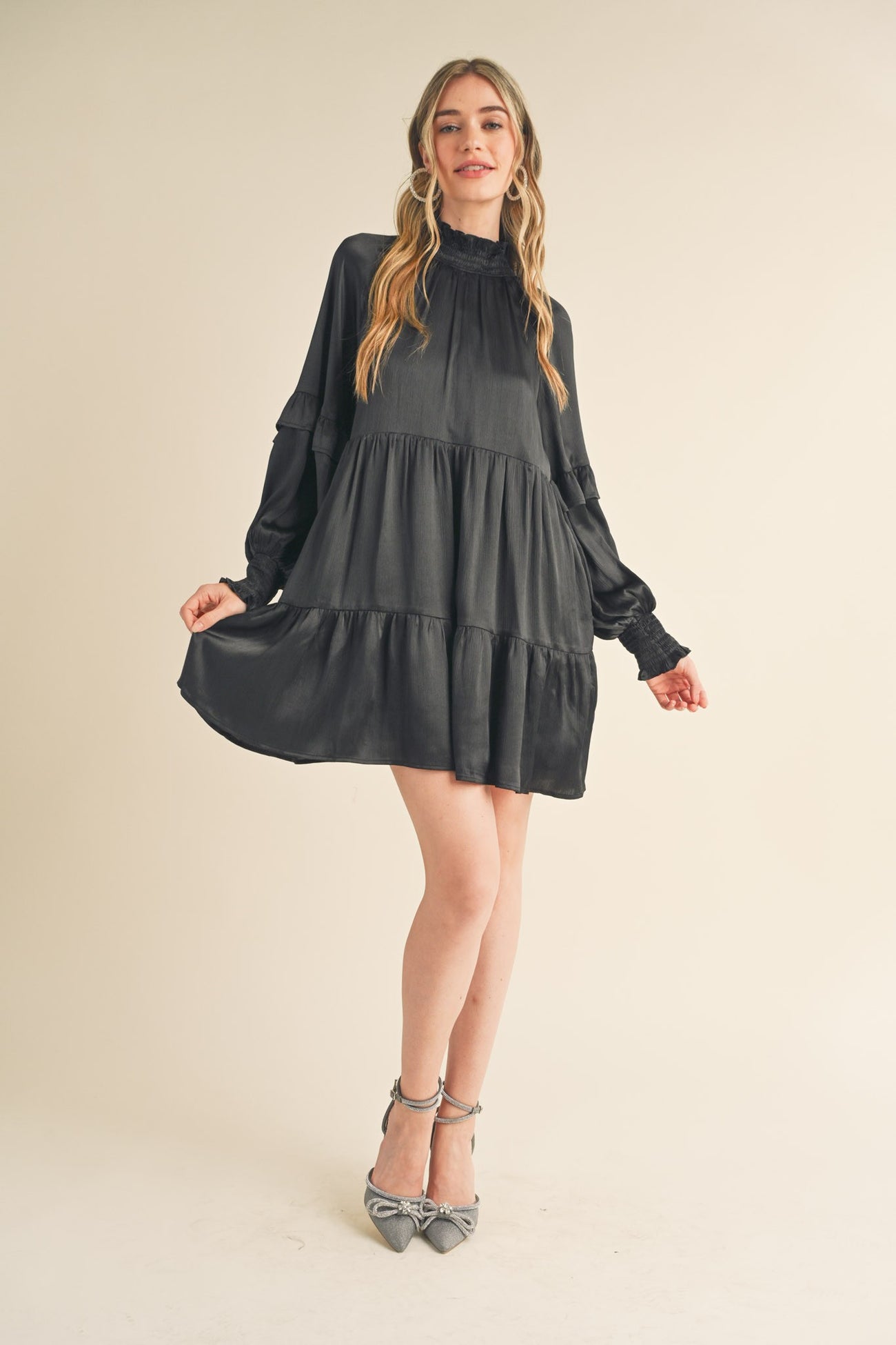 woman wearing a black tiered mini dress with long sleeves and a high neckline, paired with gray ankle strap heels