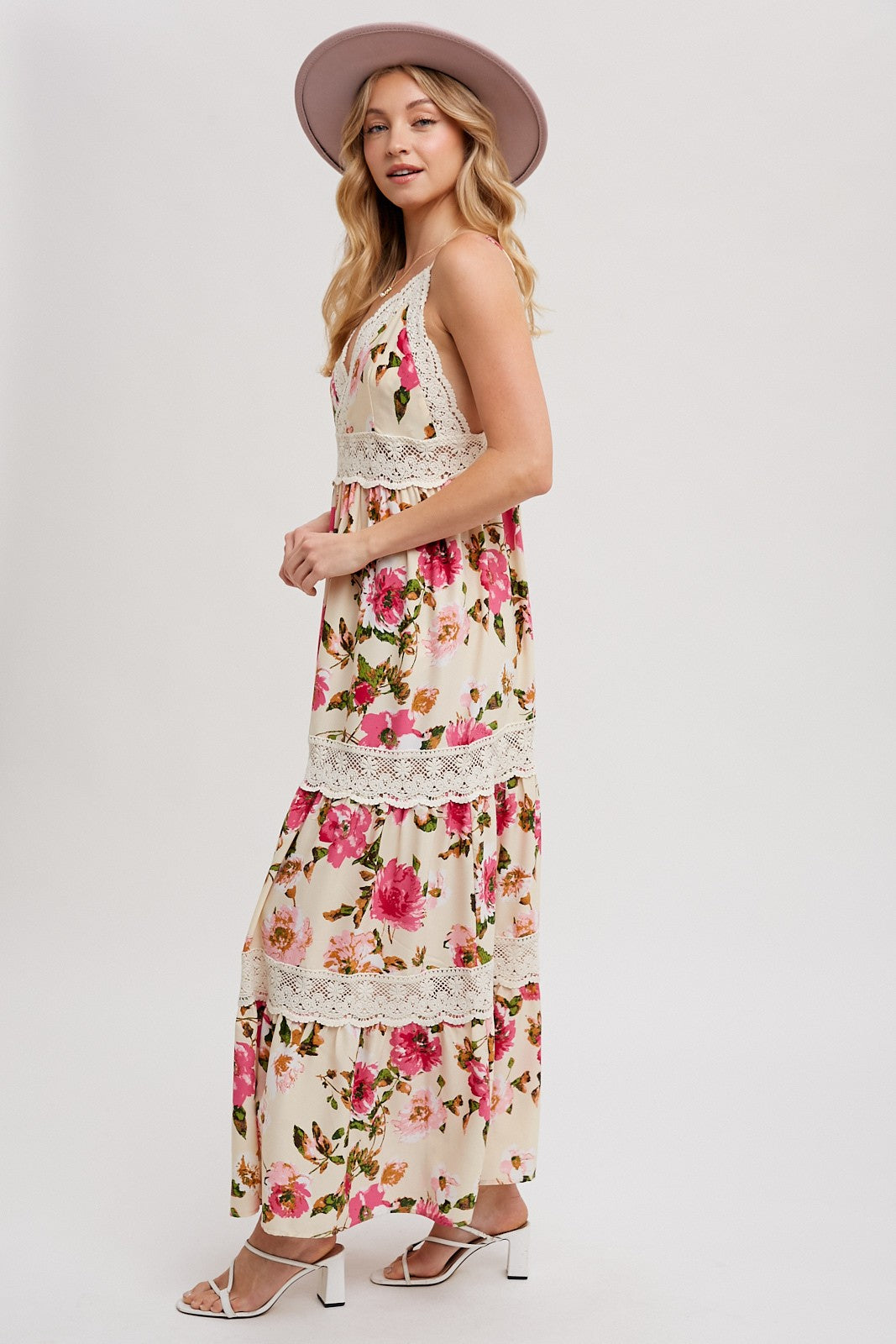 woman wearing a floral maxi dress with lace details and a wide-brimmed hat, standing sideways against a light background