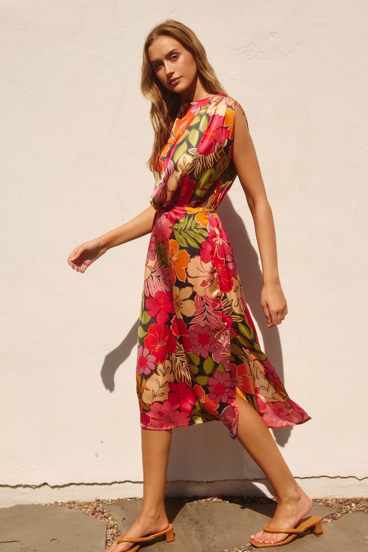 woman wearing a colorful floral midi dress with a tie waist, walking against a white wall